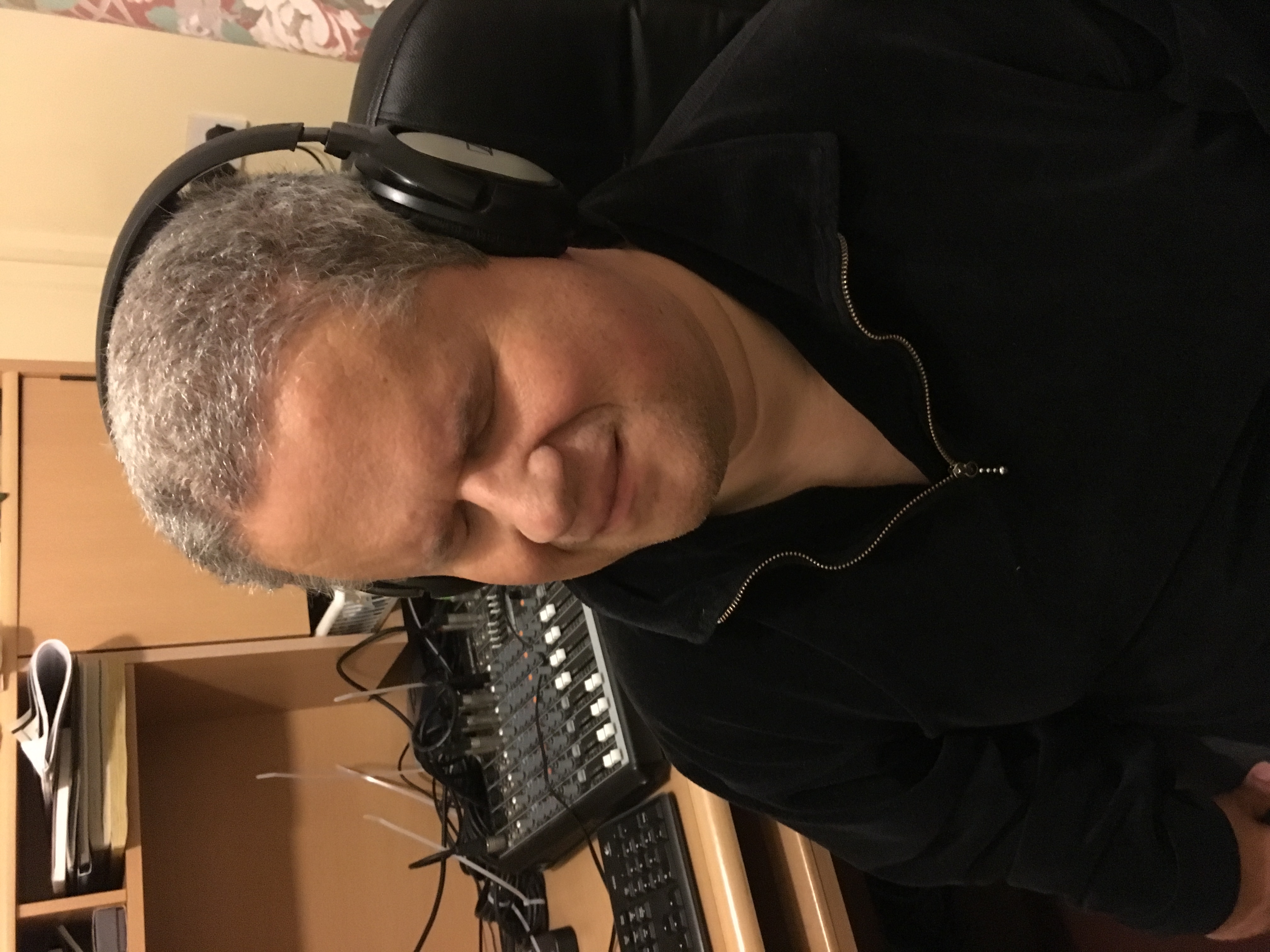 Brian has short grey hair wearing over-ear headphones. He is seated in front of a desk with a mixing console.